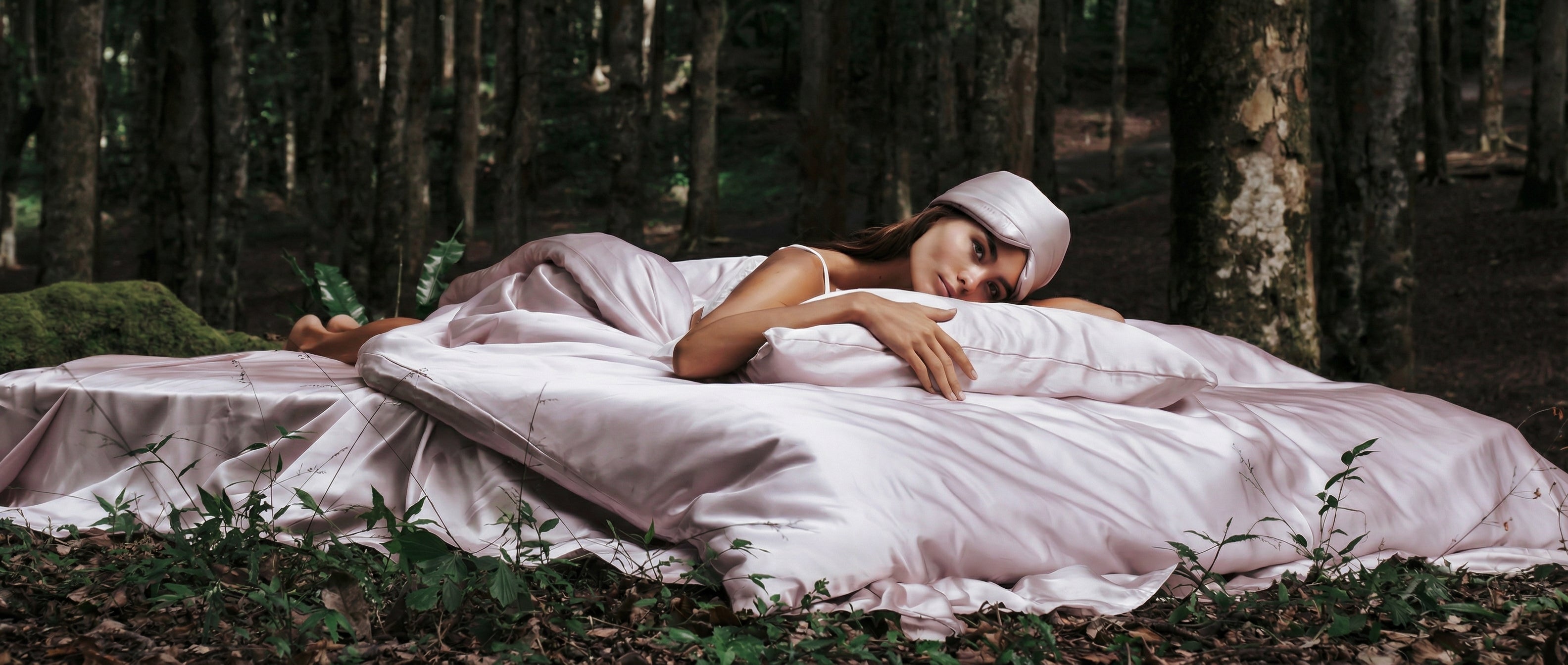A woman in a white dress lies on a bed with pale green sheets, set outdoors in a lush, green forest. She rests her head on a pillow and looks ahead peacefully.