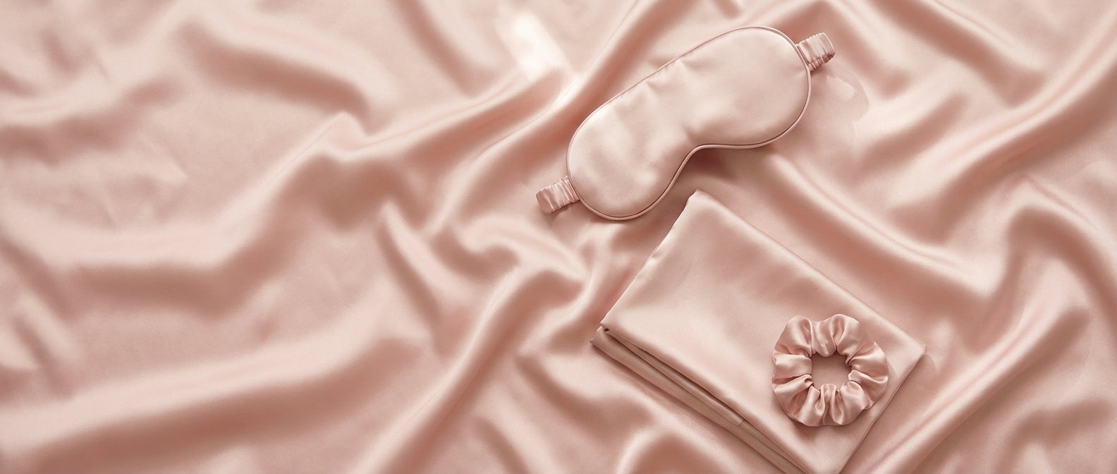 A beauty sleep gift set with a pink silk eye mask, pillowcase, and scrunchie displayed in front of a green box labeled The Beauty Sleep Gift Set.