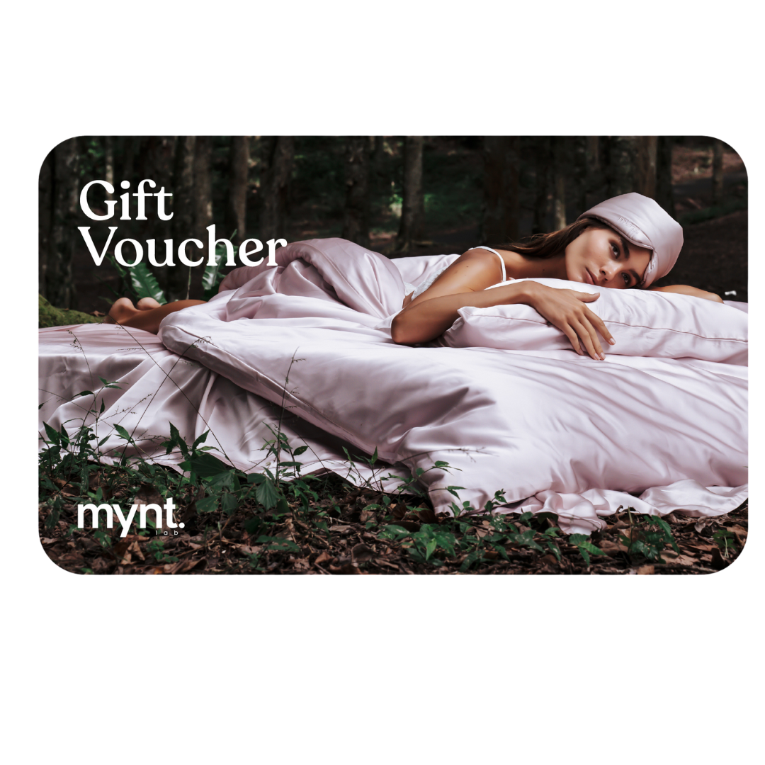 A woman in light pink sleepwear and a matching head wrap lies on a bed with pink sheets outdoors in a forest. Text reads: Gift Voucher and mynt.