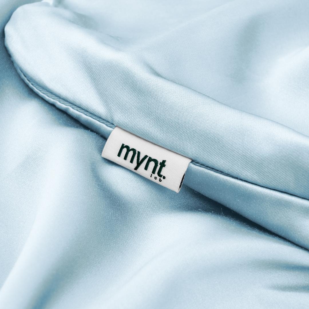 A close-up of the Fitted Sheet Set in light blue, featuring silky smooth, shiny fabric with a white "mynt lab" tag in dark green text sewn into the seam.