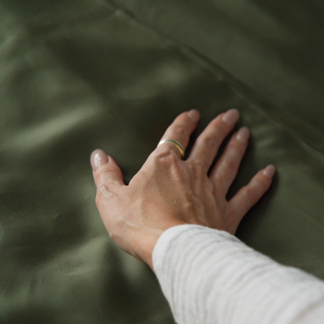 A hand with a simple ring gently rests on the silky-smooth, olive green Fitted Sheet Set. The light-coloured sleeve covers the wrist as the luxurious sheet feels soft and gentle to the touch.
