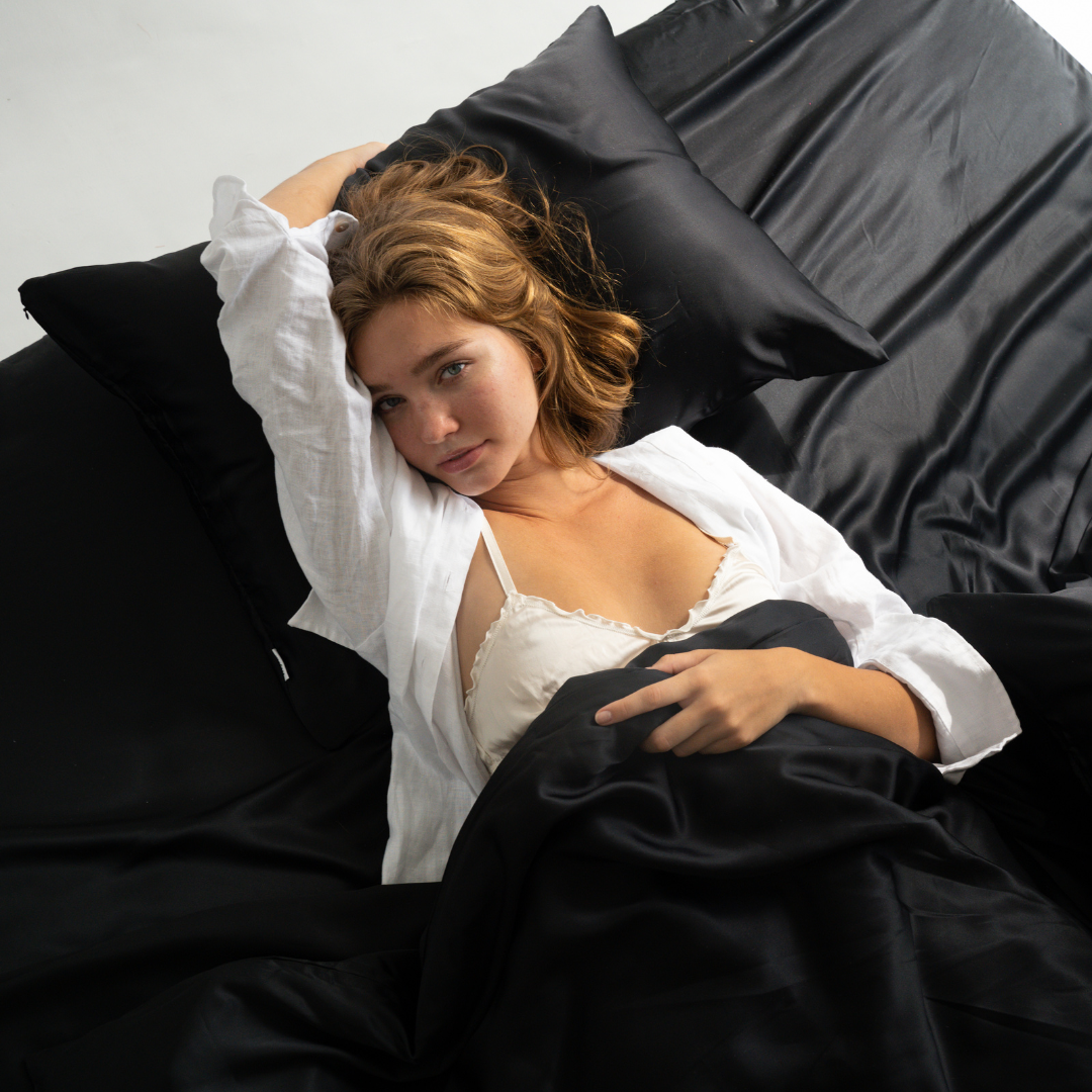 A woman with light brown hair reclines on bed dressed in the Complete Sheet Set (4-Piece) in black, wearing a white top and shirt, her arm above her head as she gazes softly at the camera.