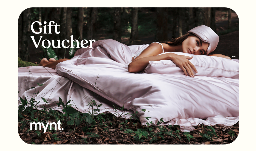 A woman in silk sleepwear and a headband lies on a bed with pink satin sheets in a forest. The text "Mynt Lab Gift Card" and "mynt." appears in white font.
