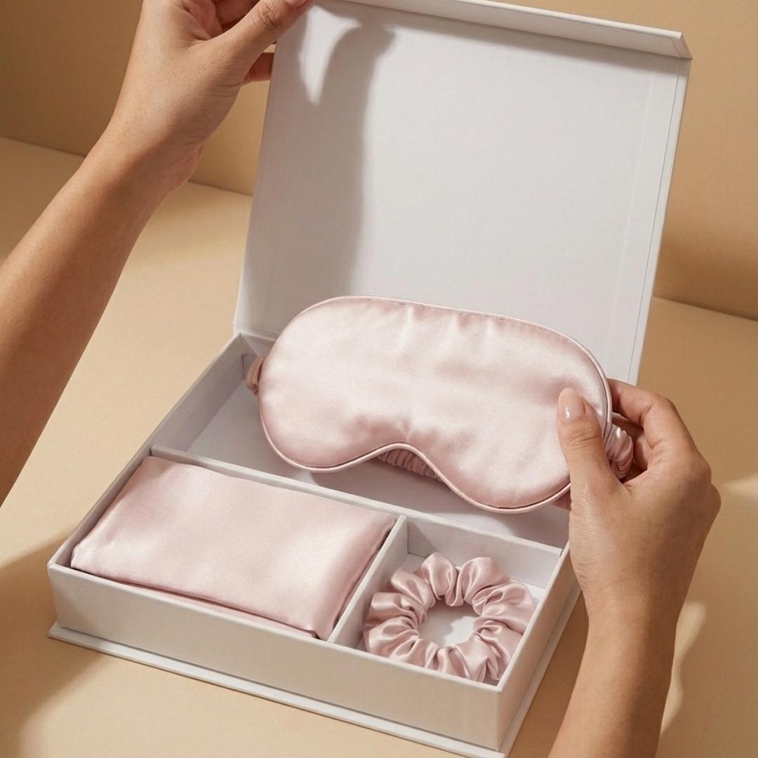 A person holds a silky pink sleep mask from a gift box set that also contains a matching scrunchie and pouch, all set against a neutral beige background.