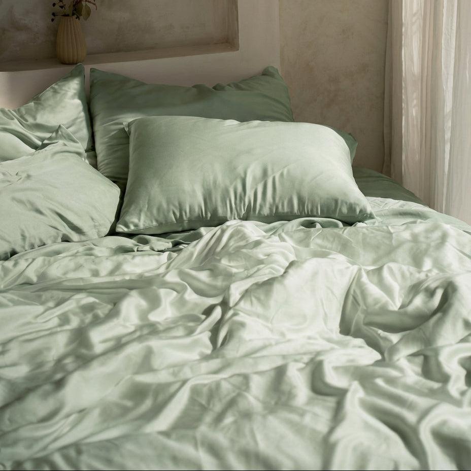 A bed dressed in light green MyntFusion™ Complete Bedding Bundle (5-Piece) by Mynt Lab is softly lit by natural light from a nearby window, with a vase of flowers placed on a recessed shelf above the pillows.