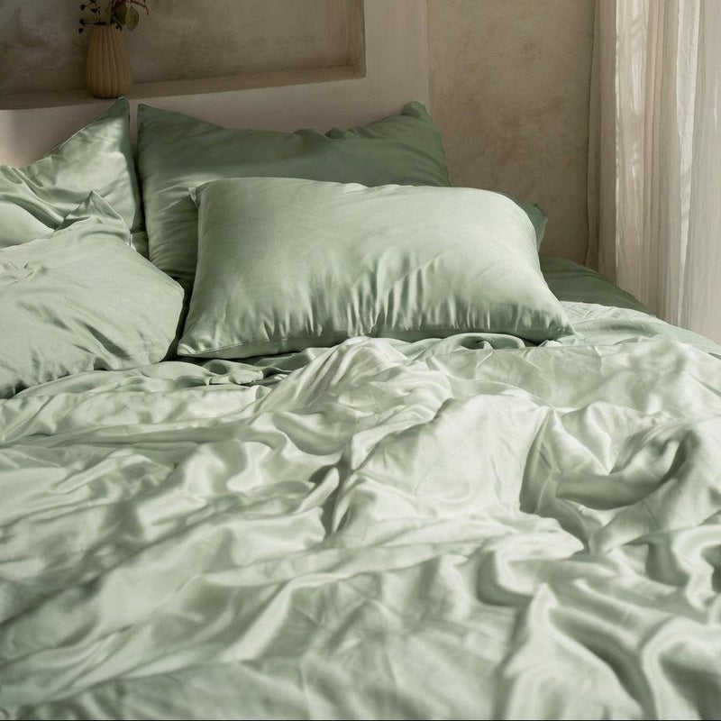 A bed dressed in light green MyntFusion™ Complete Bedding Bundle (5-Piece) by Mynt Lab is softly lit by natural light from a nearby window, with a vase of flowers placed on a recessed shelf above the pillows.