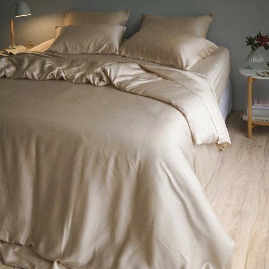 A neatly made bed with the Mynt Lab MyntFusion™ Complete Bedding Bundle (5-Piece) in light beige, complemented by pillows, sits next to a small nightstand with a lamp, vase of flowers, and book on a light wooden floor.