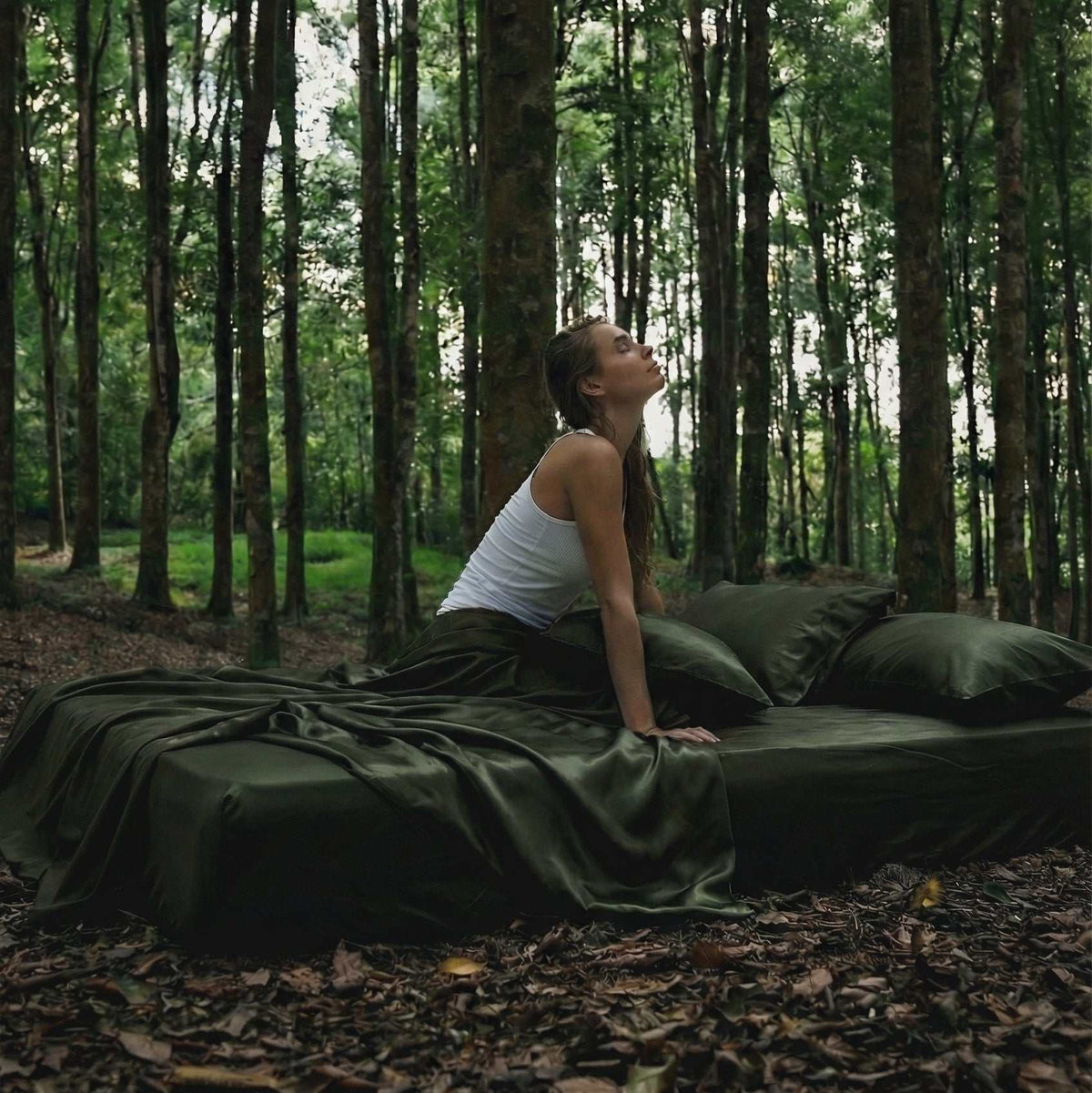 A woman in a white tank top sits on a bed with Mynt Lab's MyntFusion™ Complete Sheet Set (4-Piece), placed outdoors in a lush forest, gazing upward thoughtfully.