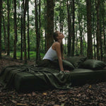 A woman in a white tank top sits on a bed with Mynt Lab's MyntFusion™ Complete Sheet Set (4-Piece), placed outdoors in a lush forest, gazing upward thoughtfully.