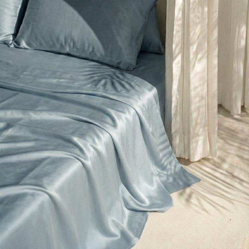 The Mynt Lab MyntFusion™ Complete Sheet Set (4-Piece) in light blue is arranged on a bed beside sheer cream curtains, with soft natural light and plant shadows creating a calm, serene space - ideal for hot sleepers.