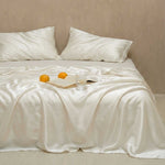 A neatly made bed features the Mynt Lab MyntFusion™ Complete Sheet Set (4-Piece) in white, perfect for hot sleepers, with two pillows. Three oranges, an open book, a glass of water, and glasses rest atop the blanket; the background wall is beige.