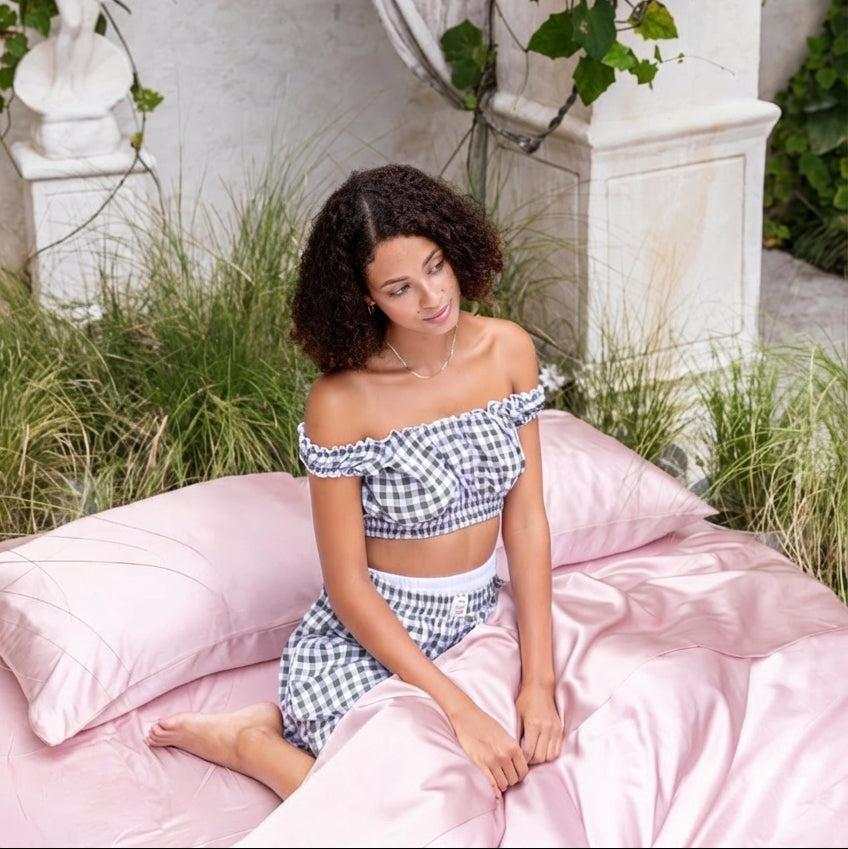 A woman with curly hair sits on a bed covered with the Mynt Lab MyntFusion™ Complete Sheet Set (4-Piece) outdoors, wearing a blue and white checkered off-shoulder top and matching skirt, surrounded by greenery and stone columns.