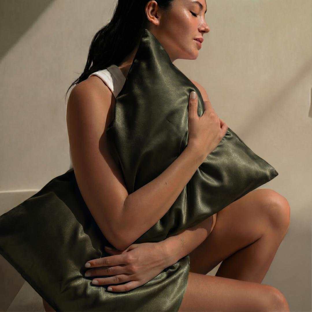 A woman with dark hair sits peacefully, eyes closed, hugging a shiny dark green pillow from the Deep Sleep Bundle, bathed in soft natural light as if drifting into deep sleep.