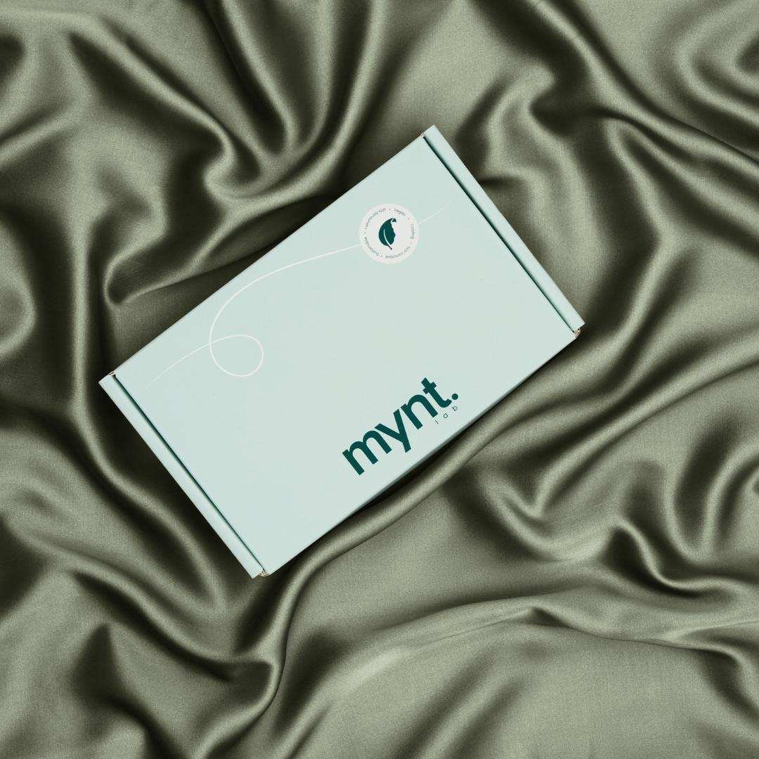 A mint green rectangular box rests on smooth olive green silky fabric. It features a small circular seal in the top right corner and an elegant white swirl design, making it perfect as a deep sleep gift set.