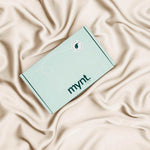 A mint green cardboard box with "mynt lab" printed on it rests on soft beige silky fabric, making the Deep Sleep Bundle - a blackout sleep mask and cooling pillowcase - an ideal gift for enhancing restful sleep.