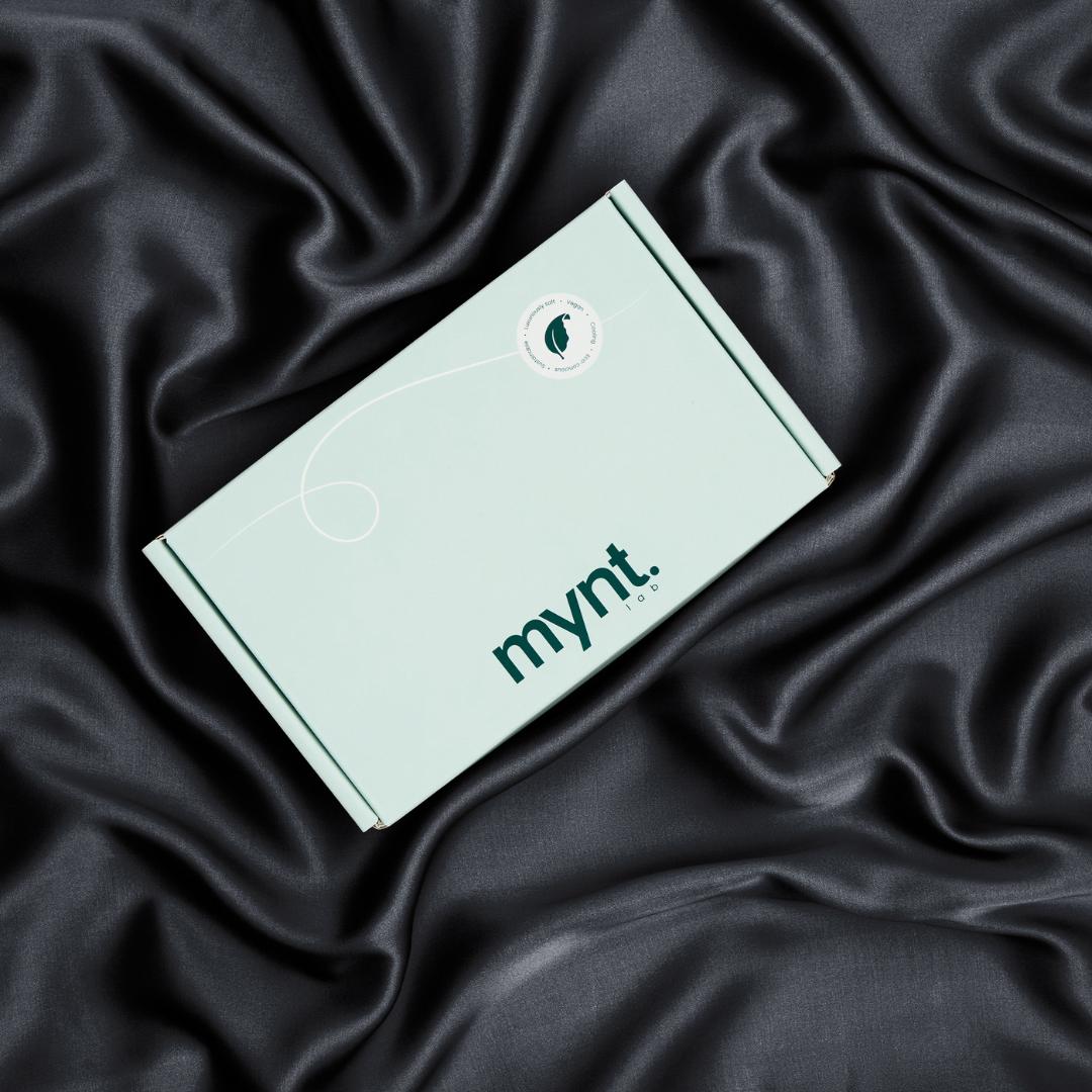 A mint green box with the mynt lab logo rests on black silky fabric. Ideal for a cooling pillowcase or blackout sleep mask,.