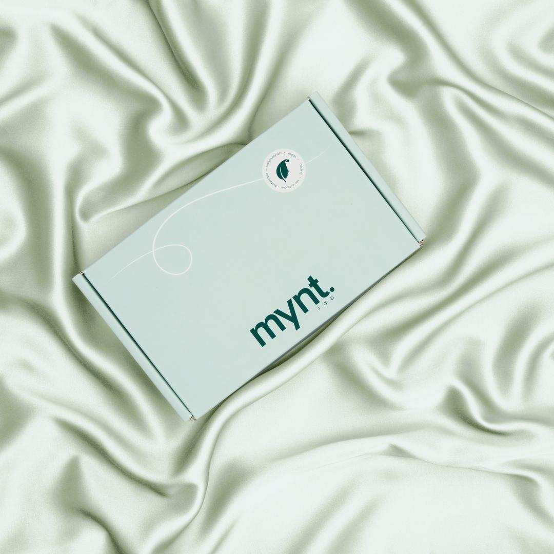 A mint green rectangular box with the mynt lab logo rests on pale green, silky wrinkled fabric resembling a luxurious cooling pillowcase, and has a white circular sticker near one corner.