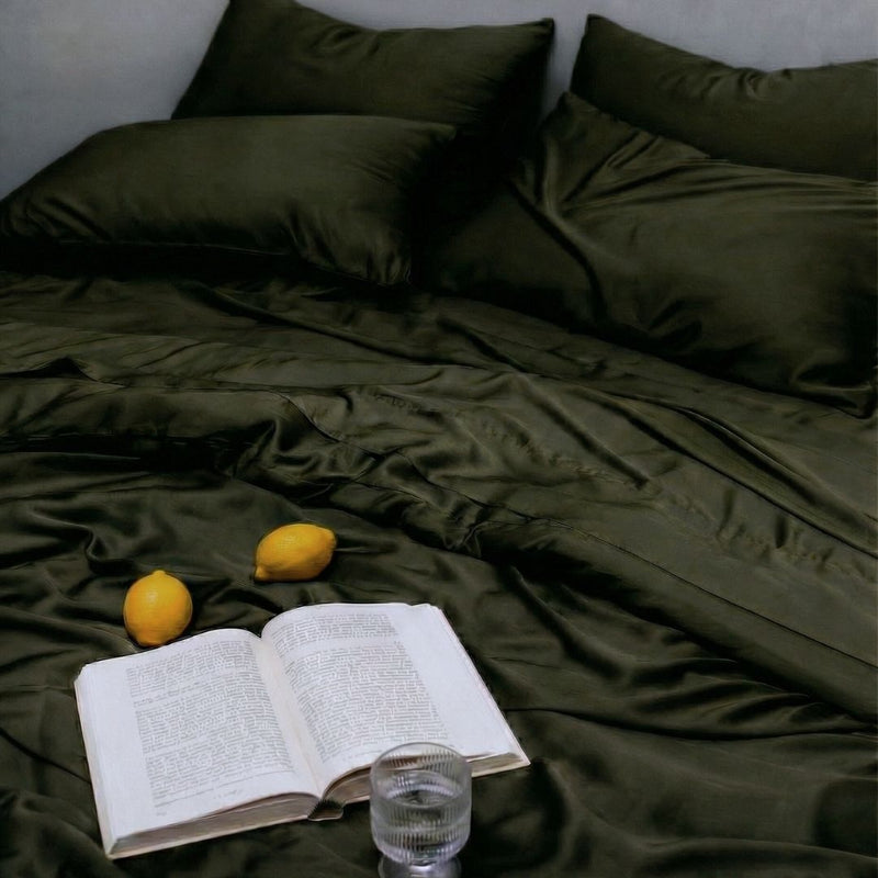 An open book, two lemons, and a glass of water sit on dark green MyntFusion™ Complete Bedding Bundle by Mynt Lab, with matching pillows visible in the background of an unmade bed.