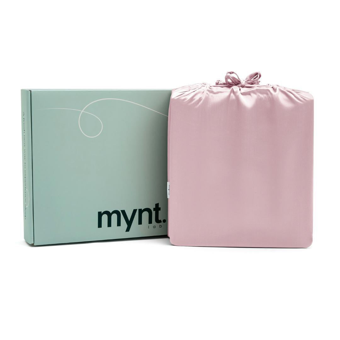 The Mynt Lab MyntFusion™ Complete Sheet Set (4-Piece) for hot sleepers, in folded pink satin, comes in a drawstring bag beside a light green box with "mynt." printed in black on the front.