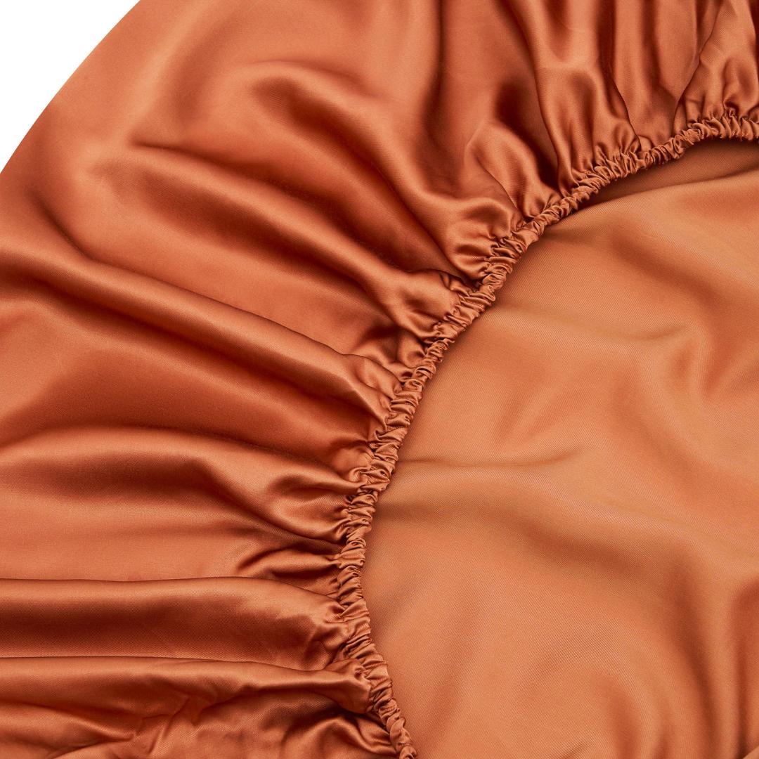 A close-up of the rust-colored MyntFusion™ Fitted Sheet Set (California King) by Mynt Lab highlights its smooth, shiny fabric and elastic edges with pleats—ideal for deep pocket mattresses.