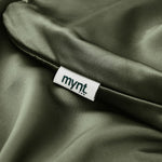 A close-up of smooth, olive-green satin on the Mynt Lab Pillowcase—perfect for beauty sleep—shows a white mynt tag in lowercase black letters, partially tucked into the seam.