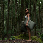 A woman stands barefoot on mossy ground in a forest, holding two large Mynt Lab Pillowcase pillows - ideal for beauty sleep - while wearing a white top, surrounded by tall trees and lush greenery.