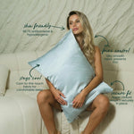 A woman sits on a cream sofa hugging a large, light blue MyntFusion™ Pillowcase by Mynt Lab, crafted from silky-smooth fabric. Text highlights its benefits: skin-friendly, reduces frizz, stays cool, and features a secure zipper closure.