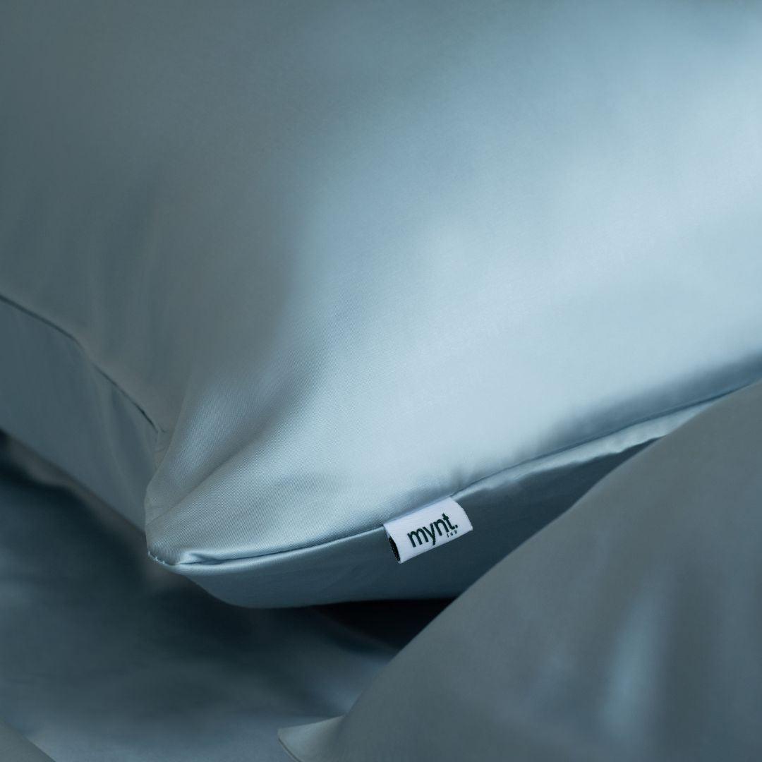 A close-up of a smooth, light blue Mynt Lab Pillowcase features a small white tag reading "mynt" in lowercase. Designed to enhance beauty sleep, the pillowcase rests on matching bedding.