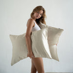 A woman in a white tank top and underwear holds two large Mynt Lab Pillowcase sand-coloured silky pillowcases, known to help reduce frizz and perfect for beauty sleep, while standing against a plain white background.