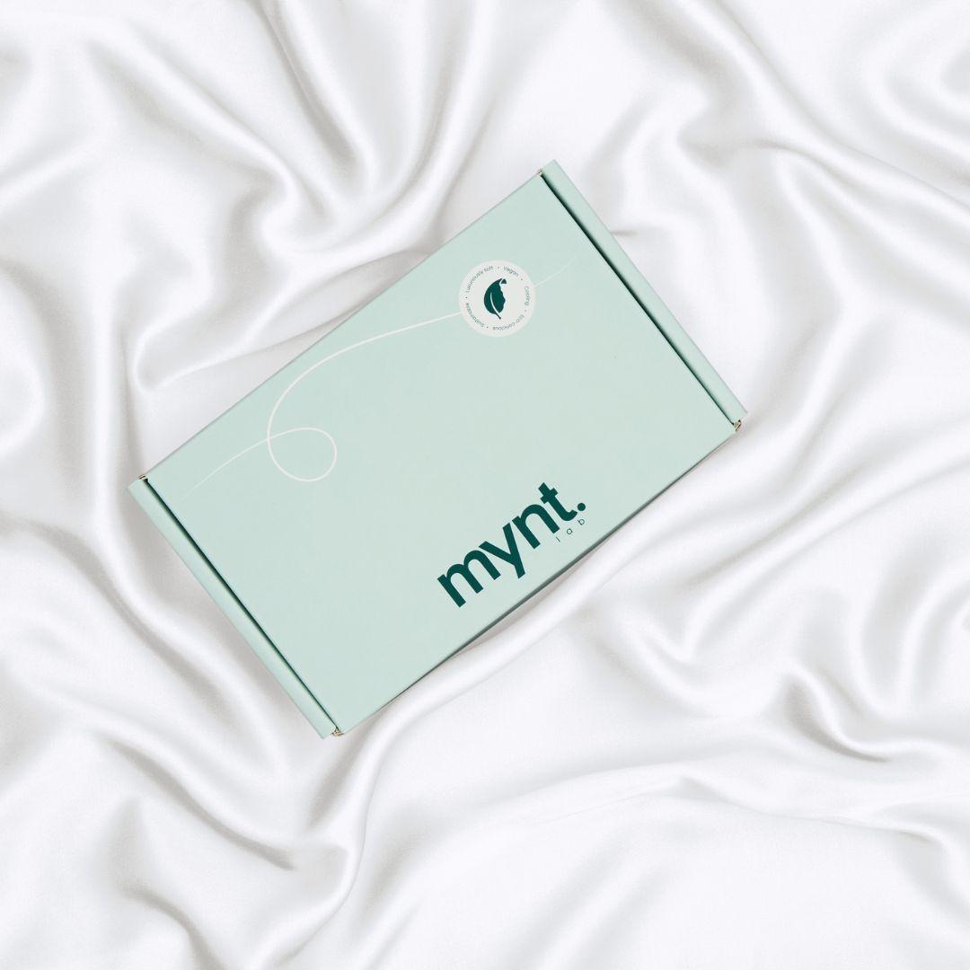 A mint green rectangular box with the mynt lab logo on the lid rests on smooth, white, silky fabric - a Pillowcase designed to enhance your beauty sleep.