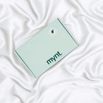 A mint green rectangular box with the mynt lab logo on the lid rests on smooth, white, silky fabric - a Pillowcase designed to enhance your beauty sleep.