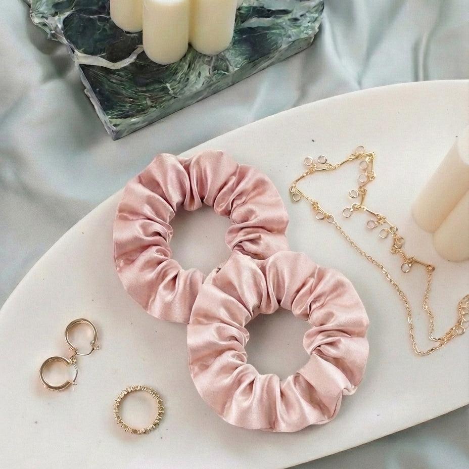 Two pink MyntFusion™ Scrunchies by Mynt Lab, crafted to be gentle on hair and reduce breakage, rest on a white tray near gold hoop earrings, a gemmed ring, and a dainty gold necklace, with candles and a marble tray in the background.