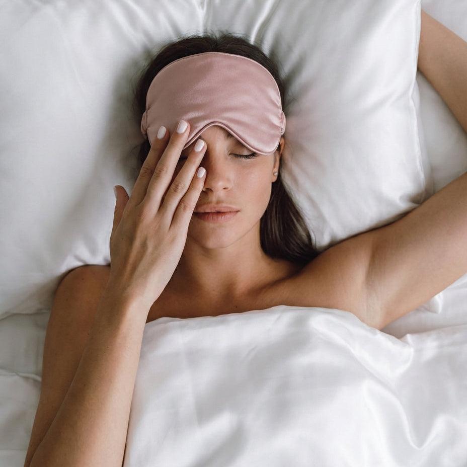 Woman wearing a pink sleep mask in bed with white pillows and sheets.