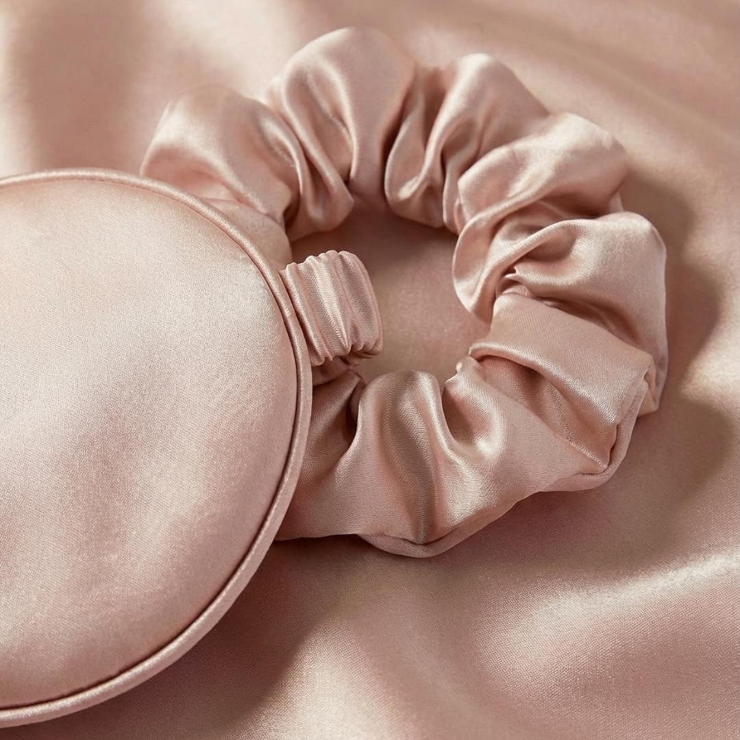 A pale pink satin scrunchie and matching sleep mask are placed on a smooth satin fabric, all in the same soft, shimmery color.