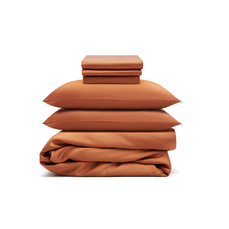 The Bedding Bundle, featuring rust-colored antibacterial sheets, pillowcases, two pillows, and a comforter, is neatly stacked and folded against a plain white background.