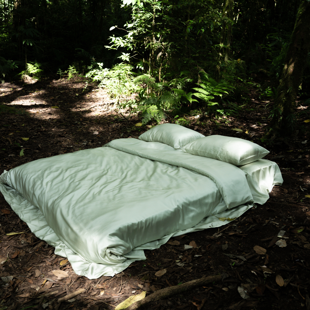 A Bedding Bundle with light green, antibacterial bedding and pillows is arranged neatly outdoors on a forest floor, surrounded by lush green plants and trees in natural sunlight.