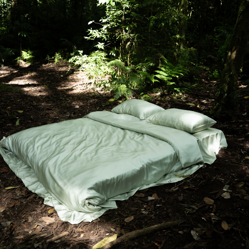 A Bedding Bundle with light green, antibacterial bedding and pillows is arranged neatly outdoors on a forest floor, surrounded by lush green plants and trees in natural sunlight.