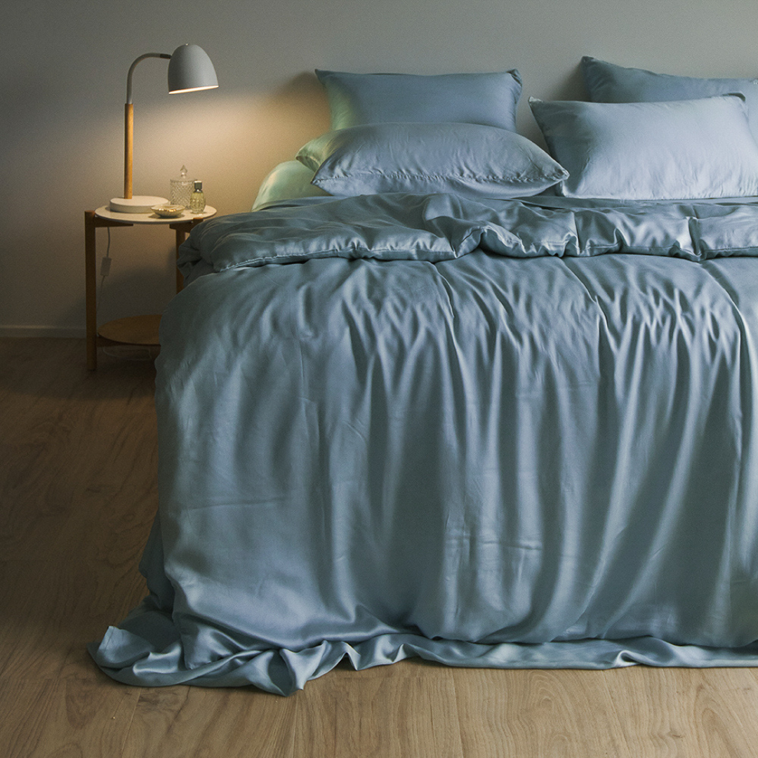 A neatly made bed featuring the light blue Bedding Bundle and matching pillows sits by a small wooden nightstand with a lamp, glass of water, and book on light wood flooring in a cozy, softly lit room.