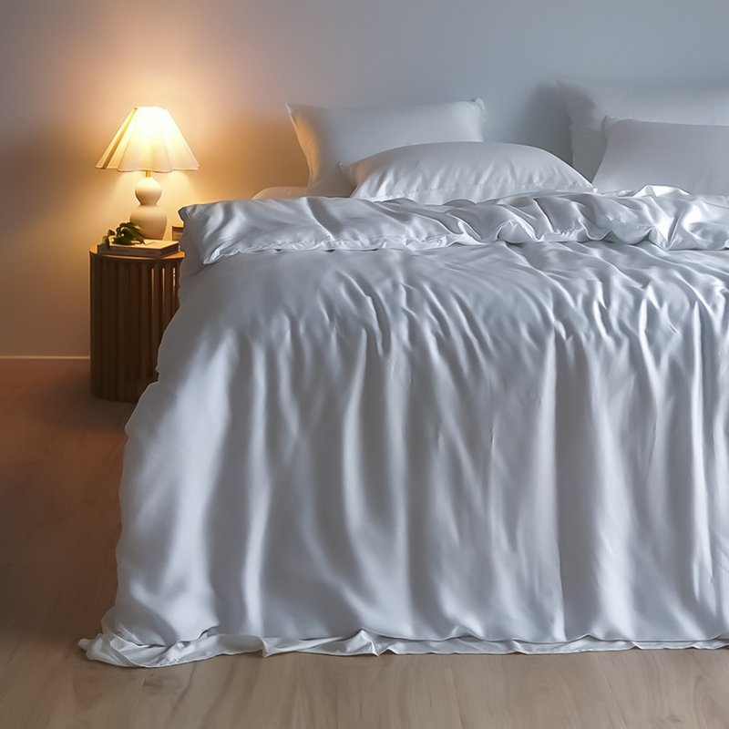 A neatly made bed features the Bedding Bundle—customisable white satin sheets and pillows—next to a wooden nightstand with a lit lamp and decor, in a softly lit bedroom with light-colored flooring.