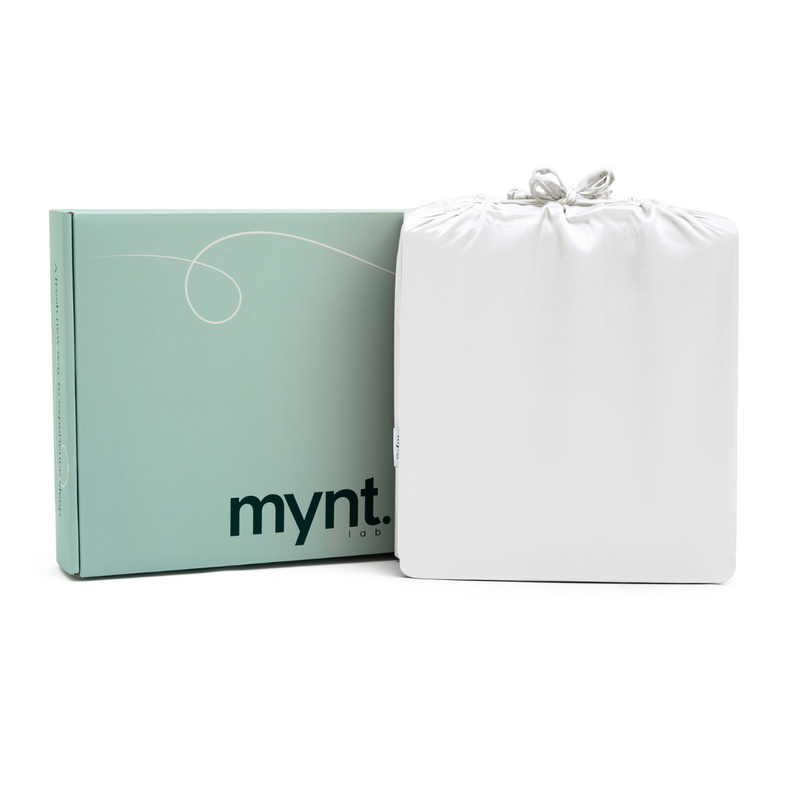 A folded white Fitted Sheet Set in a silky drawstring bag is placed beside a light green mynt lab box with black text, both displayed against a white background.