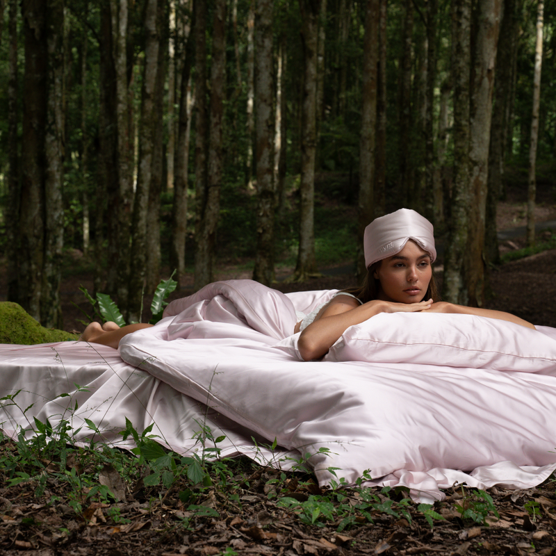 A woman rests on a large, silky pink Bedding Bundle in a forest, her pale headscarf blending with the customizable bedding and natural beauty of tall trees and lush greenery.