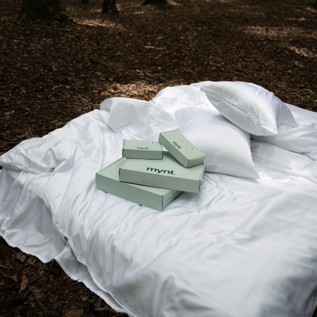 A white satin bedspread and pillow rest on a forest floor, with several light green boxes labeled mynt—each containing the Bedding Bundle for cooling comfort—arranged on the bedspread under natural daylight.