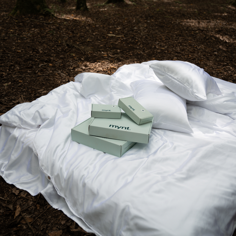 A white satin bedspread and pillow rest on a forest floor, with several light green boxes labeled mynt—each containing the Bedding Bundle for cooling comfort—arranged on the bedspread under natural daylight.
