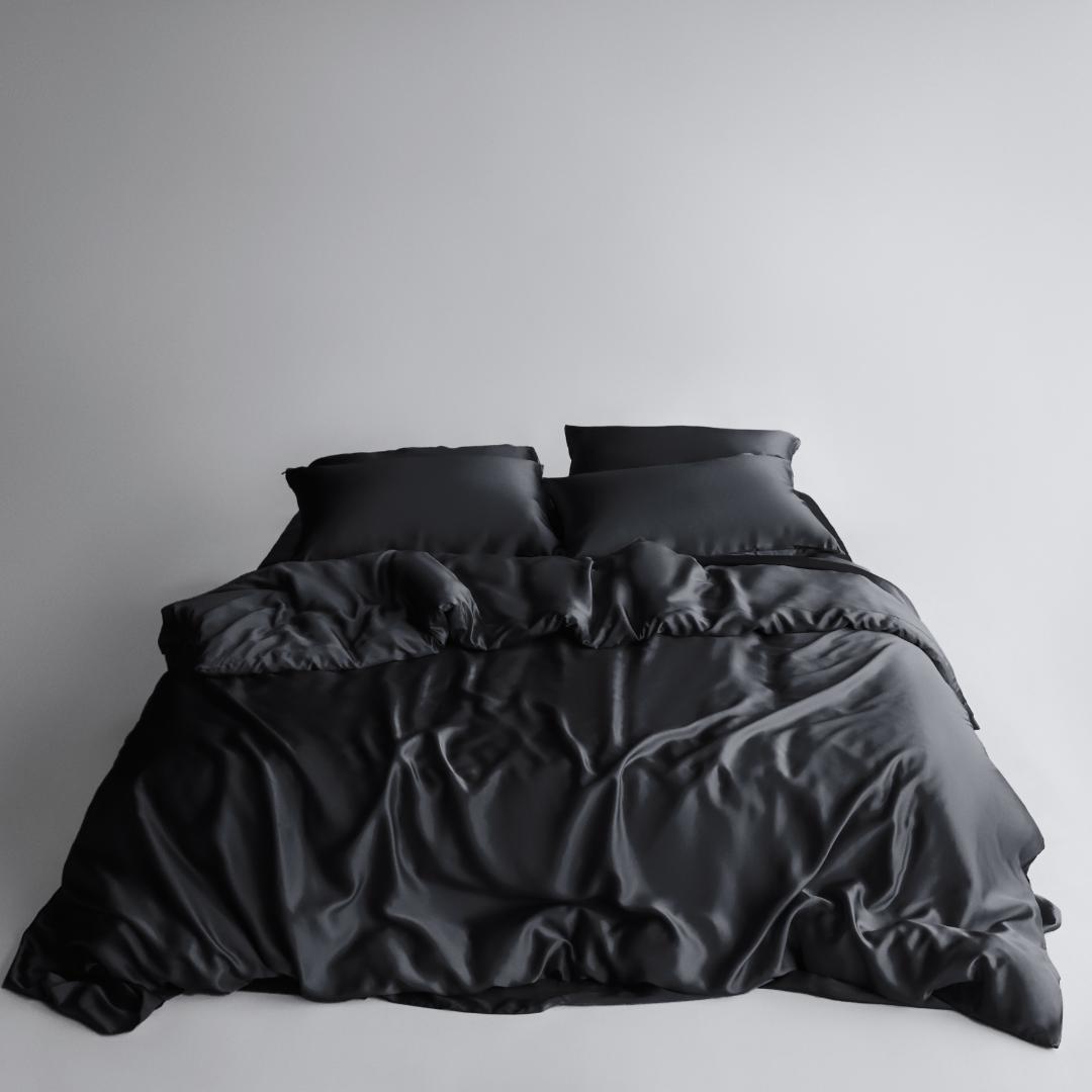A neatly made bed features the Bedding Bundle in black silky, cooling fabric with two pillows, set against a plain light background. The smooth, shiny bedding shows slight wrinkles and folds for a relaxed look.