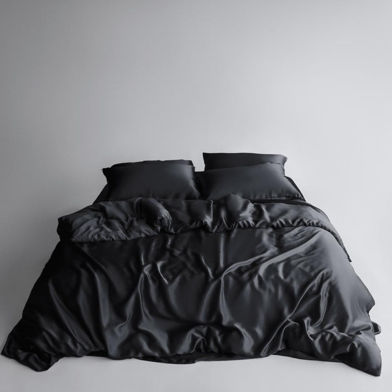 A neatly made bed features the Bedding Bundle in black silky, cooling fabric with two pillows, set against a plain light background. The smooth, shiny bedding shows slight wrinkles and folds for a relaxed look.