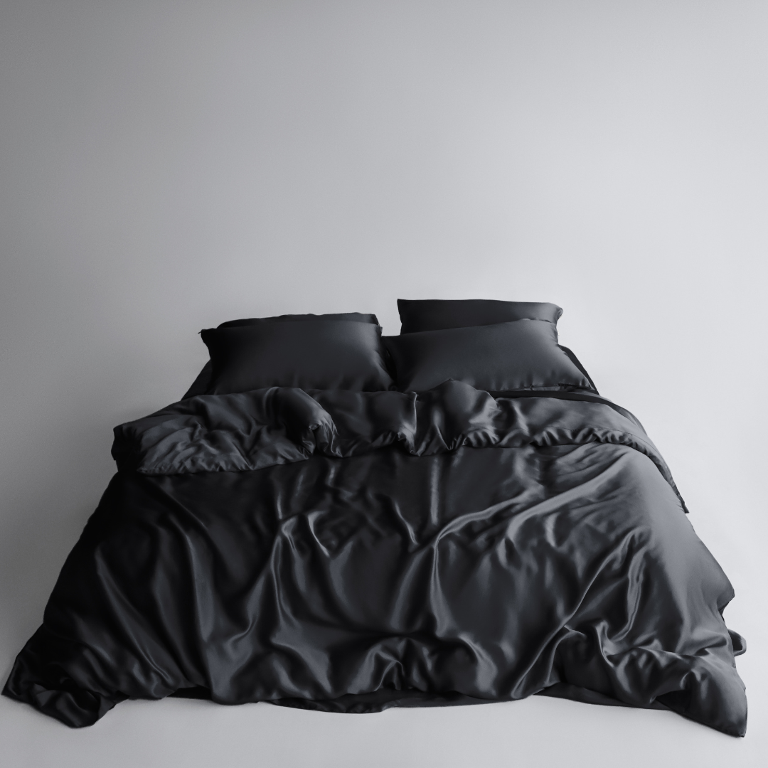 A neatly made bed features the Bedding Bundle in black silky, cooling fabric with two pillows, set against a plain light grey background. The smooth, shiny bedding shows slight wrinkles and folds for a relaxed look.
