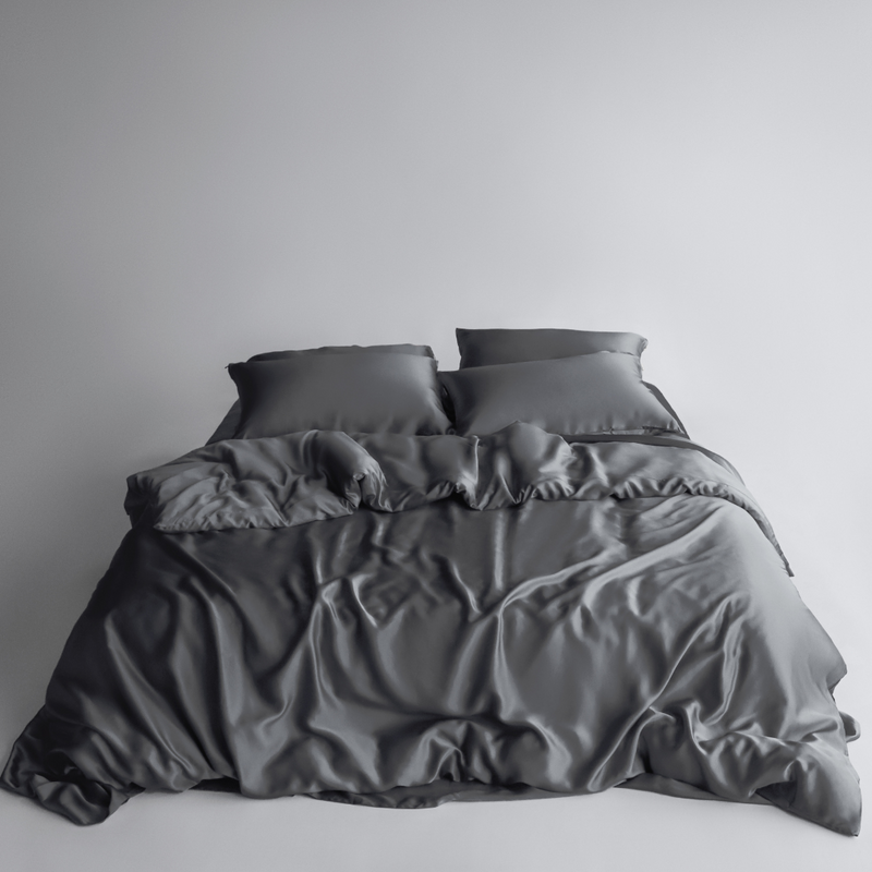 A neatly made bed with two pillows and a dark grey silky duvet cover showcases the Bedding Bundle, customisable to match your unique style and needs, set against a simple light background.