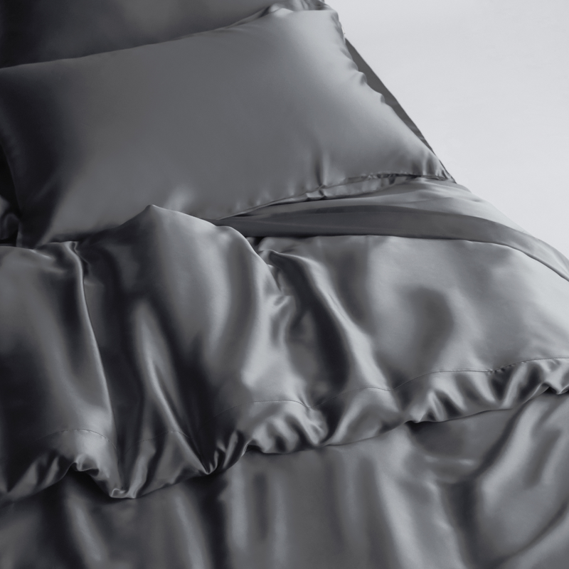 A close-up of a bed shows the Duvet Cover in smooth, shiny dark grey silky fabric with a matching pillow, highlighting its sustainable materials for a luxurious and elegant look.