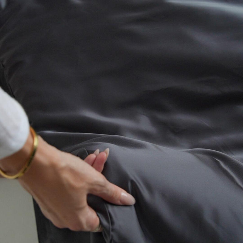 A person’s manicured hand with a gold bracelet glides over the silky, charcoal-coloured Bedding Bundle, showcasing the sheets' luxurious, smooth texture.