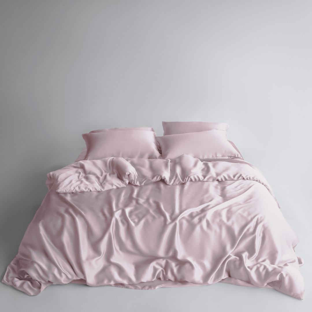 A neatly made bed features the Bedding Bundle in light pink silky fabric with two matching pillows, set against a plain white background. The slightly rumpled blanket adds a soft and inviting look.